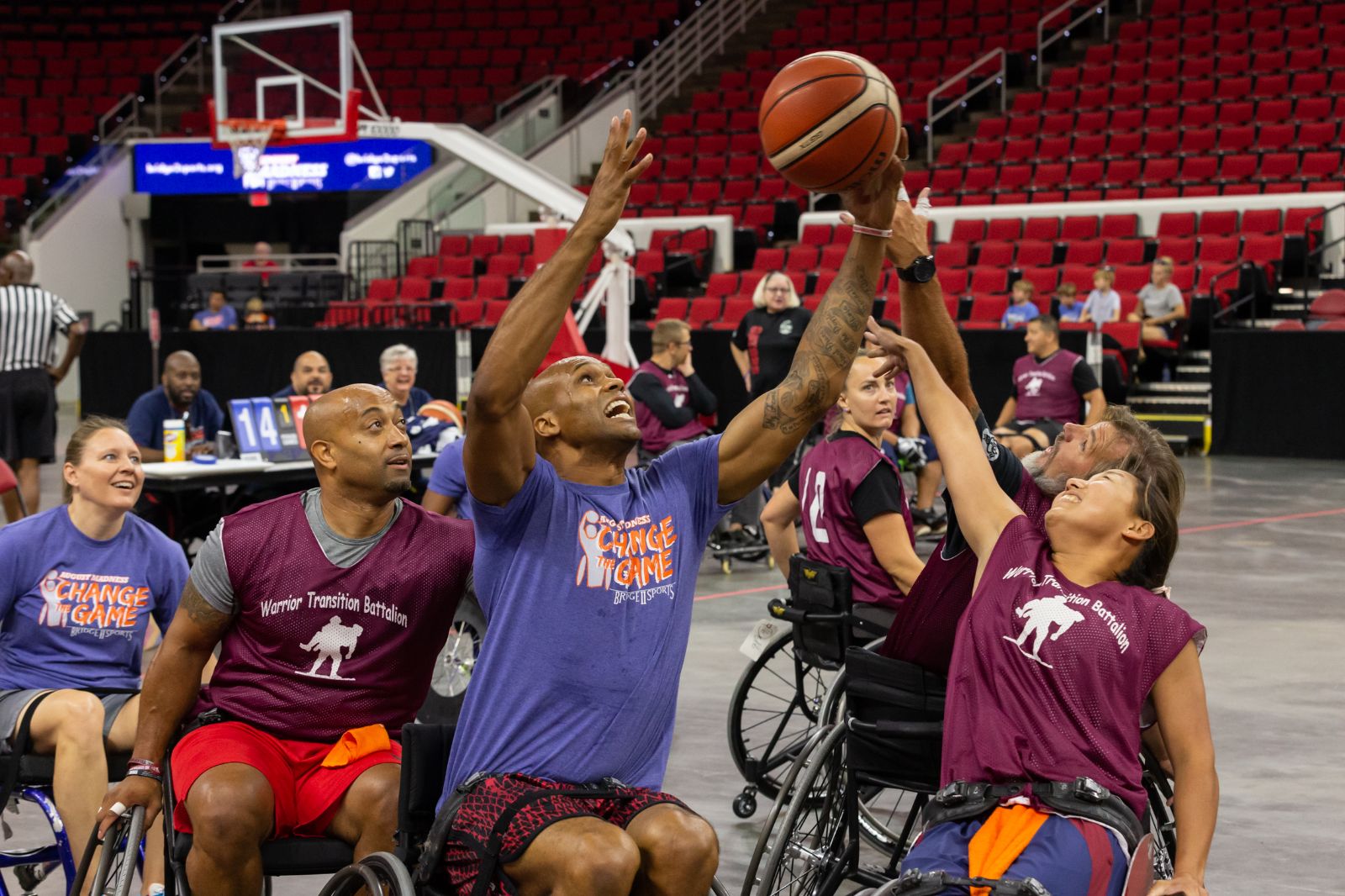 A man in a sports chair goes for a lay up while two opposing players try to block the shot.  The players are diverse in both gender and skin tone.