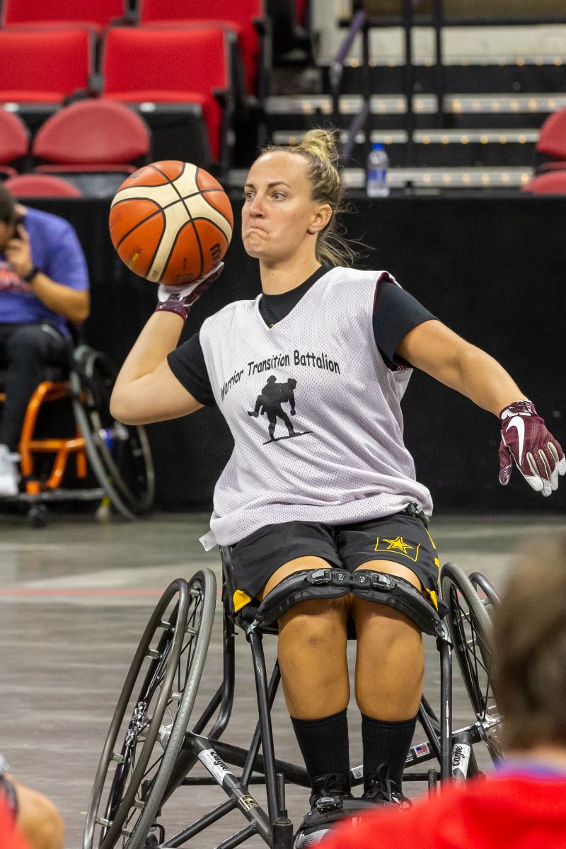 A light skinned woman in a sports chair shown in the act of beginning a basketball pass; she is wearing a shirt that says, "warrior transition battalion".