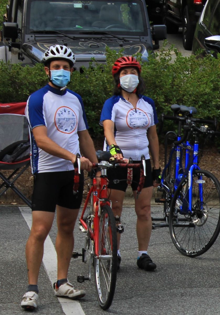 Two riders, one male in appearance one female in appearance, standing in front of a tandem cycle.  Both are wearing helmets and jerseys.