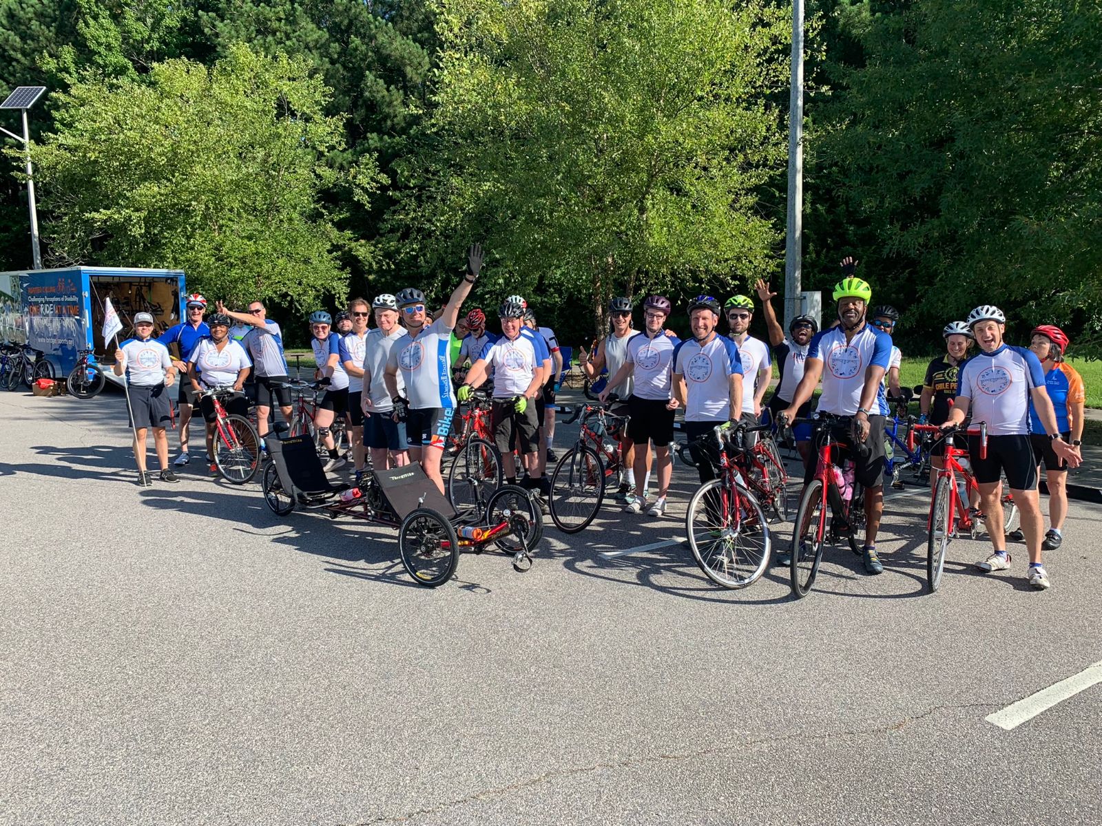 A large group of people standing together and posing for a picture.  There are cycles in front of and amongst the riders, and a trailer off to the left side.