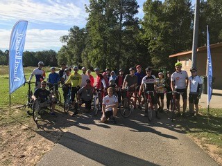 A picture of a group of cyclists posing for a picture in the foreground.  The background is a cloud streaked blue sky on the left, trees in the center, and a building on the right.