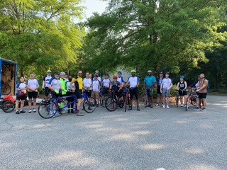 A group of cyclists posing in a parking lot in the foreground; the background consists of tall trees with a small building partially visible on the left side.