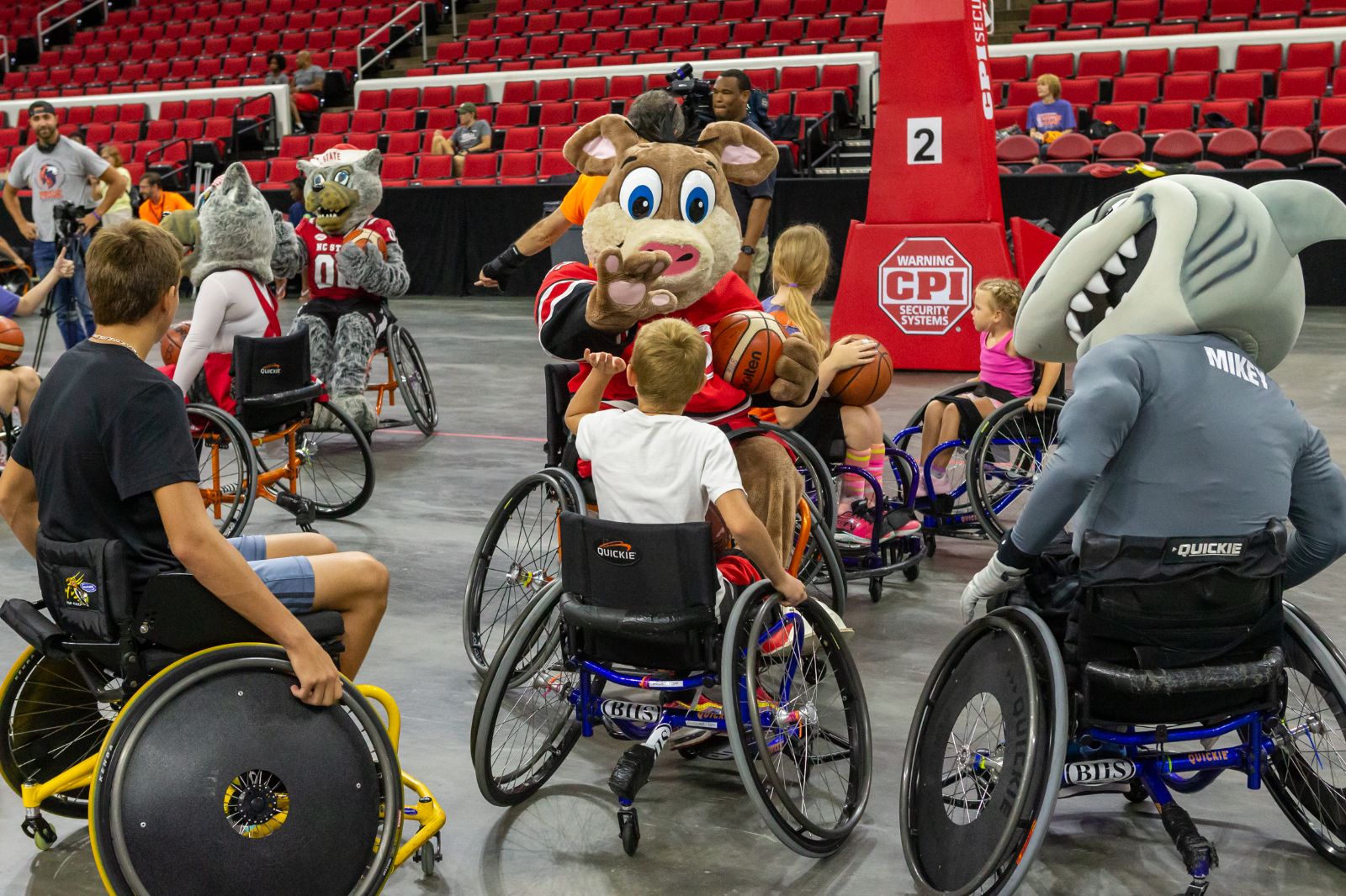 A group of young players in sports chairs interacting with other people, dressed as sports' mascots, also in sports chairs.